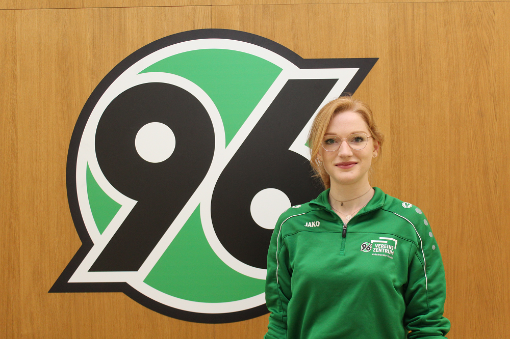 Eine Person in grüner Jacke steht vor einem großen 96-Logo an einer Holzwand.