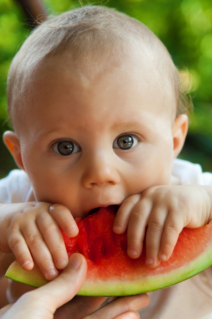 Baby hält ein Stück Wassermelone in der Hand und isst es, während es mit großen Augen nach oben schaut, im Freien.