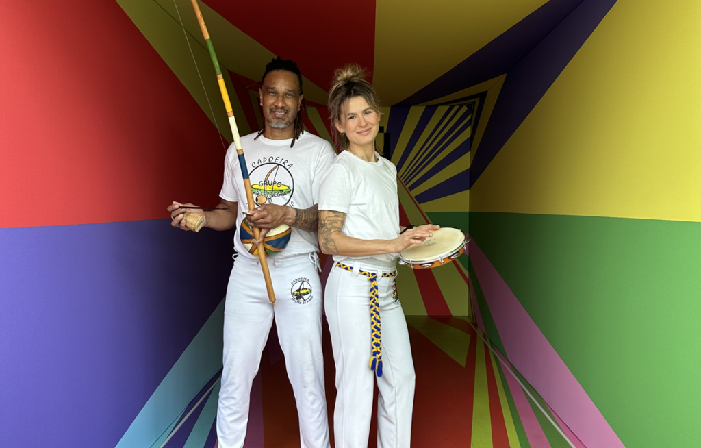 Zwei Personen in weißen Capoeira-Kostümen stehen in einem bunten, geometrischen Flur und halten Capoeira-Instrumente.