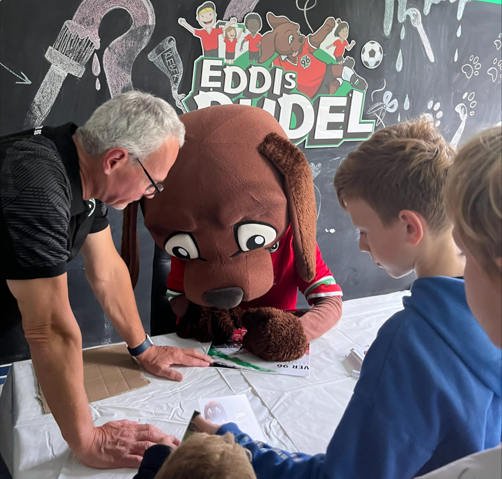 Eine Person in einem Hundemaskottchen-Kostüm gibt Kindern an einem Tisch Autogramme, während ein Mann assistiert.