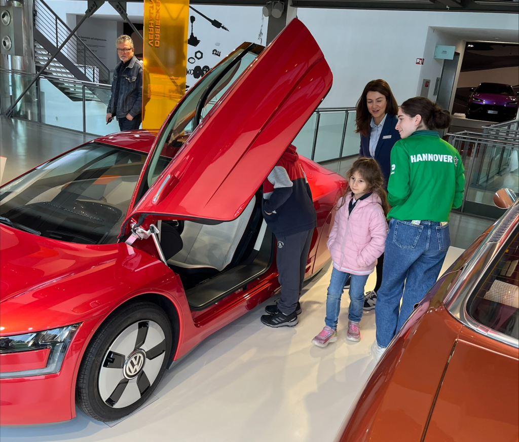 Eine Gruppe von Personen, darunter zwei Kinder, betrachtet einen roten Volkswagen mit geöffneter Fahrertür in einem Innenraum.