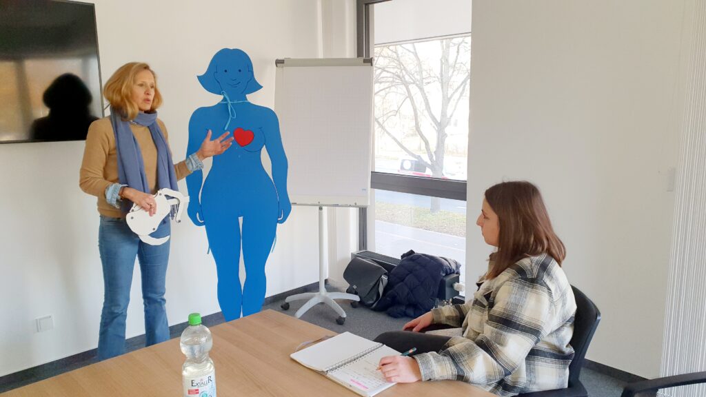 Eine Frau erklärt anhand eines Herzdiagramms auf einer blauen Figur, während eine andere Frau dasitzt und sich Notizen macht.