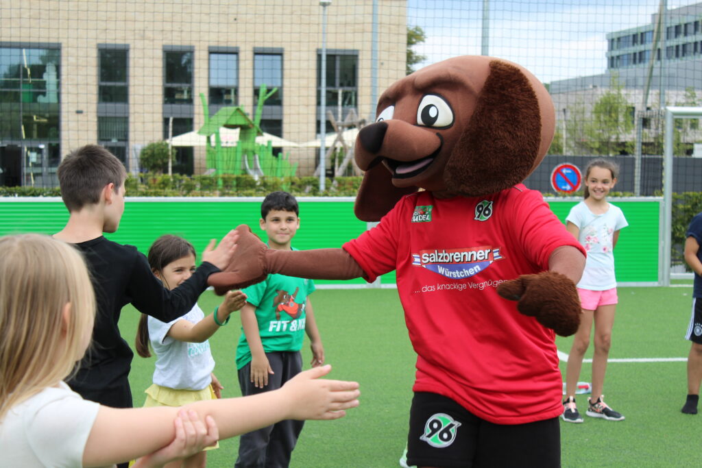 Ein Hundemaskottchen in einem roten Hemd gibt lachenden Kindern auf einem Fußballplatz ein High Five.