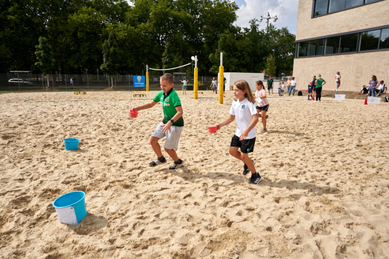 Kinder rennen auf einem Sandplatz um die Wette und tragen Becher mit Wasser zu blauen Eimern unter einem sonnigen Himmel.