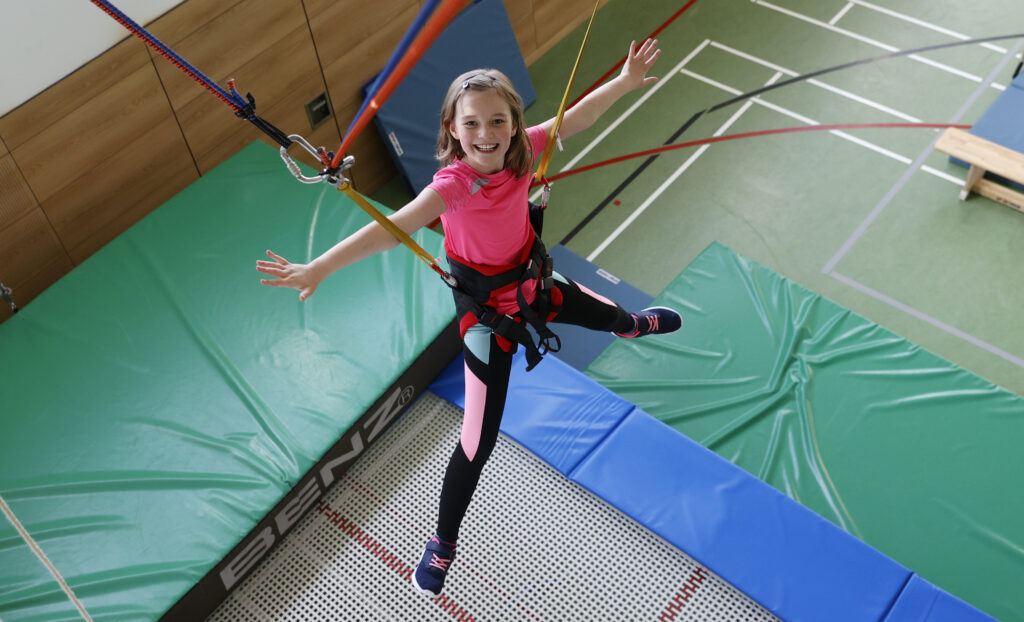 Lächelndes Mädchen in einem Gurtzeug, das mit ausgestreckten Armen auf einem Trampolin in einer Turnhalle hüpft.
