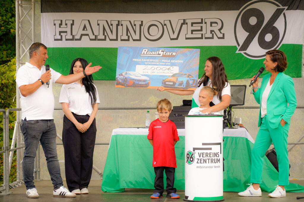 Fünf Personen und ein Kind auf der Bühne vor einer Hannover 96-Fahne, die einen großen Scheck über 2000 € in der Hand hält.