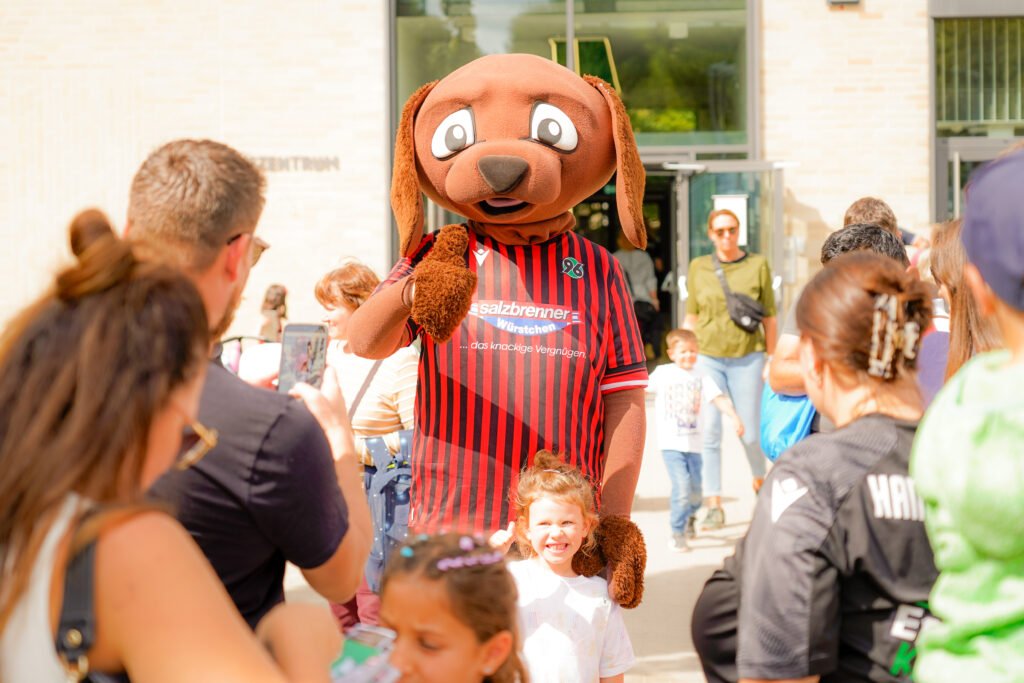 Eine Person in einem Hundemaskottchenkostüm posiert mit lächelnden Kindern und Erwachsenen im Freien an einem sonnigen Tag.