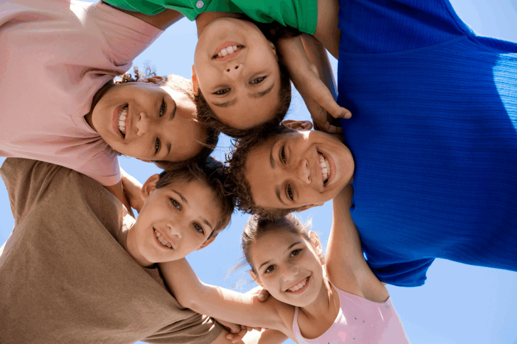 Fünf lächelnde Kinder stehen im Kreis, die Arme umeinander gelegt, und blicken vor einem blauen Himmel in die Kamera.