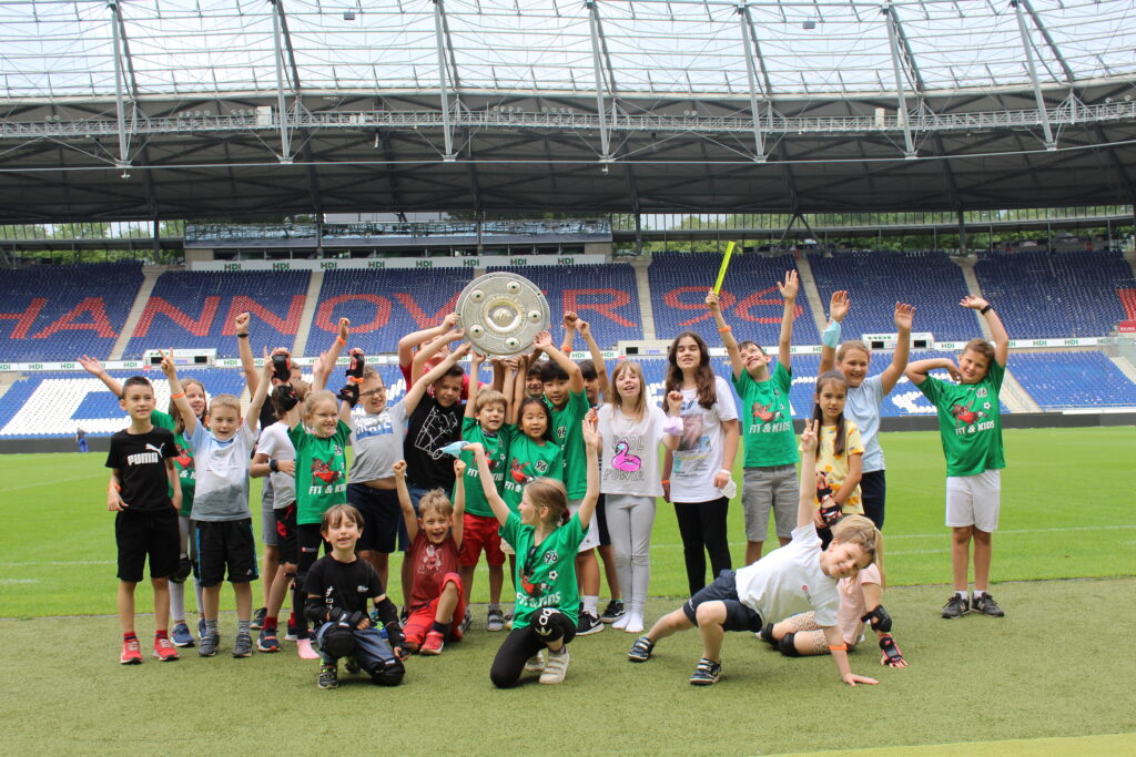 Eine Gruppe von Kindern, die auf einem Fußballplatz feiern, wobei ein Kind einen großen silbernen Pokal in der Hand hält.