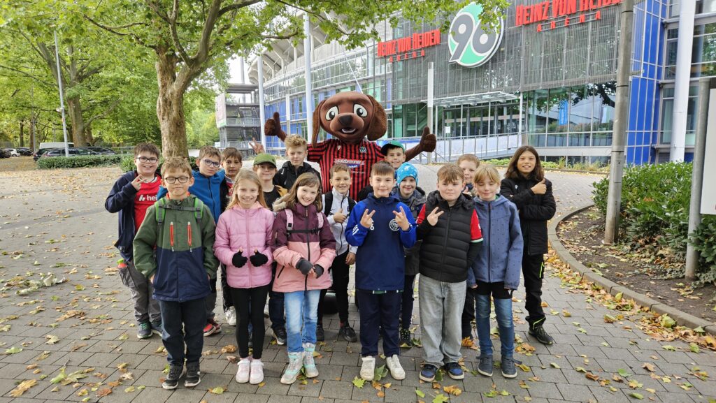 Eine Gruppe von Kindern posiert mit einem Maskottchen vor einem Stadion, das von Bäumen und Herbstblättern umgeben ist.