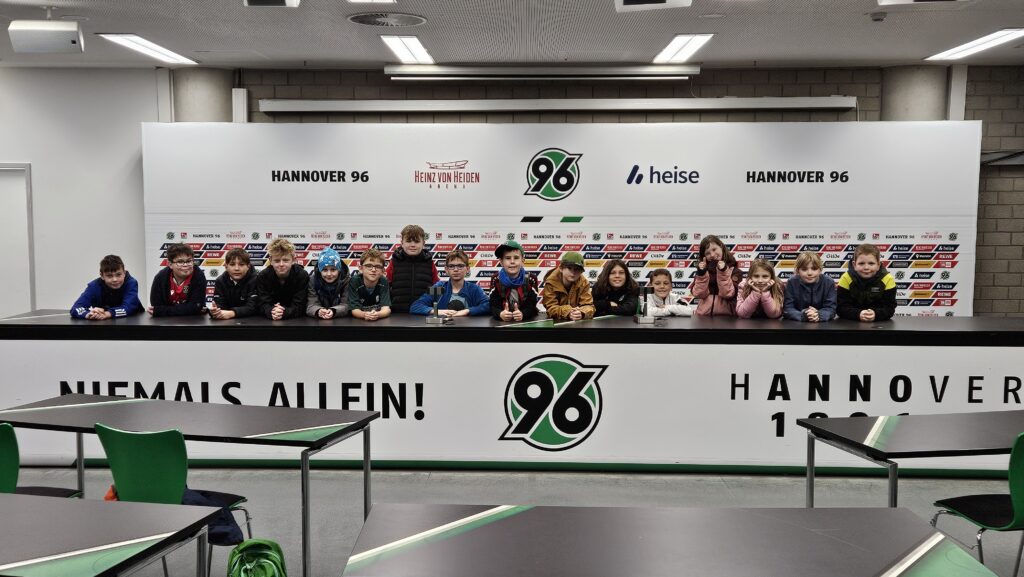Eine Gruppe von Kindern sitzt hinter dem Tisch der Pressekonferenz von Hannover 96 und lächelt in die Kamera.